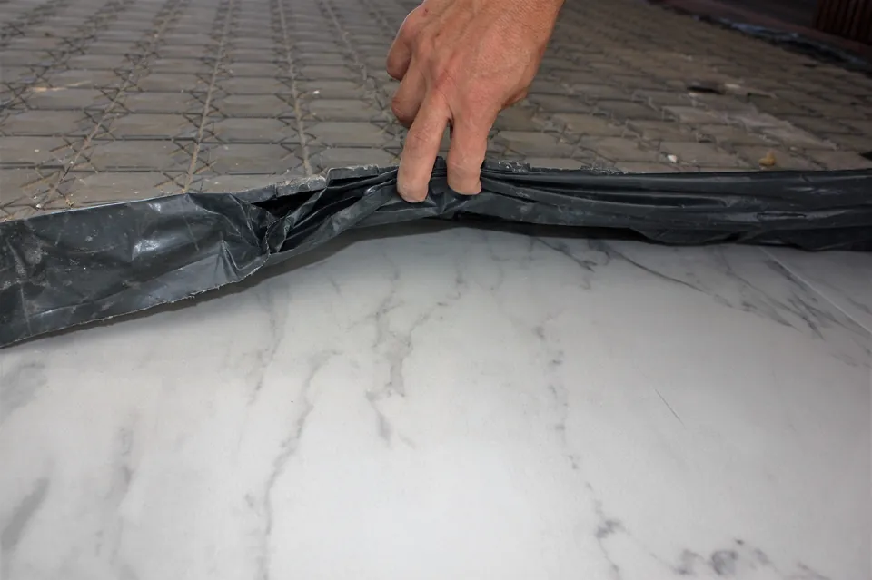 Mão levantando a borda da proteção, com as placas e a lona por cima e o piso branco de veios cinza intacto por baixo