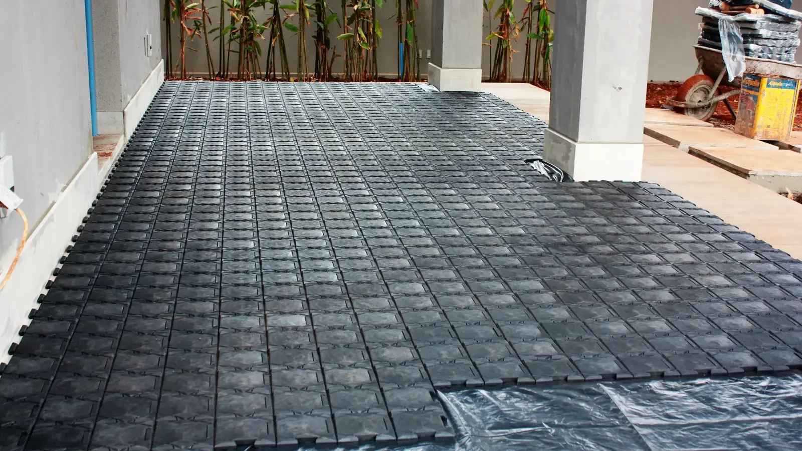 Varanda em obra com o piso coberto por placas pretas de proteção encaixadas, a lona aparecendo na borda e um carrinho de mão ao fundo